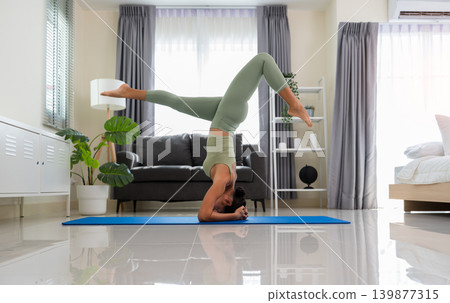 Side view of sporty woman in green top and leggings doing yoga in Salamba sirsasana exercise or Headstand pose on blue mat in cozy living room at home. Asian female doing balance and core strength Side view of sporty woman in green top and leggings doing yoga in Salamba sirsasana exercise or Headstand pose on blue mat in cozy living room at home. Asian female doing balance and core strength 139877315