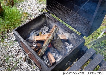A charcoal grill with burning wood and smoke rising. The grill is placed on a gravel surface surrounded by green grass. A charcoal grill with burning wood and smoke rising. The grill is placed on a gravel surface surrounded by green grass. 139877958