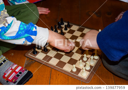 Two children playing chess on a wooden floor. One child has light skin and wears a colorful shirt. The other child has dark skin and wears a blue shirt. Two children playing chess on a wooden floor. One child has light skin and wears a colorful shirt. The other child has dark skin and wears a blue shirt. 139877959