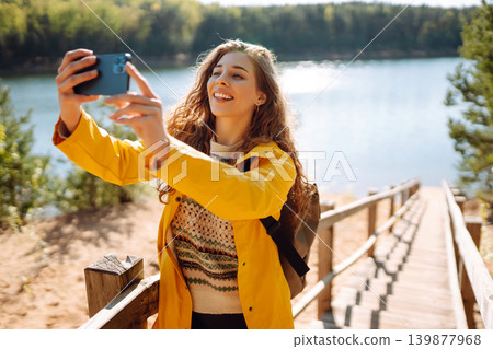 Happy woman with phone in hands taking selfie in forest. Young tourist enjoying spring day near lake 139877968