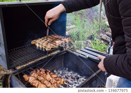 A person grilling meat on a barbecue. The scene shows skewers of chicken and vegetables cooking over hot coals. Smoke rises in a natural outdoor setting. A person grilling meat on a barbecue. The scene shows skewers of chicken and vegetables cooking over hot coals. Smoke rises in a natural outdoor setting. 139877975