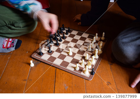Two children playing chess on a wooden floor. One child has light skin and wears colorful socks. The other child has dark skin and is focused on the game. Two children playing chess on a wooden floor. One child has light skin and wears colorful socks. The other child has dark skin and is focused on the game. 139877999