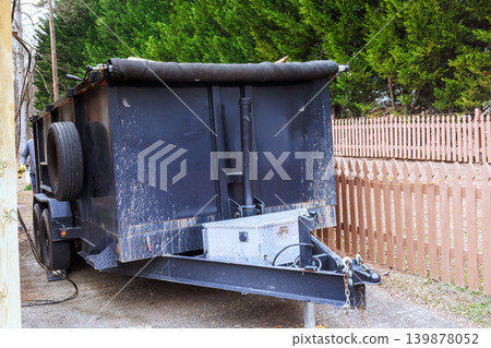 Trash trailer is parked next to fence in residential space with green trees nearby. Trash trailer is parked next to fence in residential space with green trees nearby. 139878052