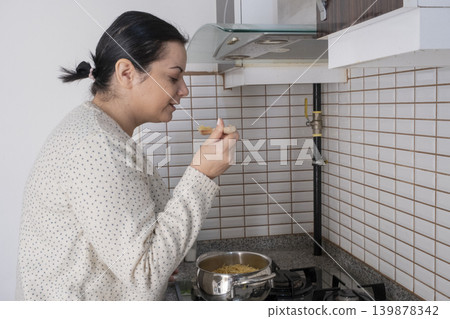 Woman Tasting Food While Cooking in Kitchen. 139878342