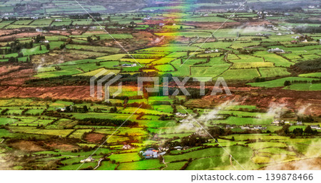 A striking rainbow descends over the vibrant patchwork of green fields and scattered white farmhouses in rural Ireland, observed from above on a clear day. A striking rainbow descends over the vibrant patchwork of green fields and scattered white farmhouses in rural Ireland, observed from above on a clear day. 139878466