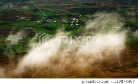 High-angle view of low clouds and mist drifting across a rural landscape of green agricultural fields and small houses. High-angle view of low clouds and mist drifting across a rural landscape of green agricultural fields and small houses. 139878467