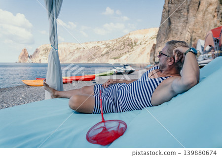 Man beach relaxation senior man in vintage striped swimwear reclining on a luxury sunbed overlooking the Mediterranean sea cliffs Man beach relaxation senior man in vintage striped swimwear reclining on a luxury sunbed overlooking the Mediterranean sea cliffs 139880674