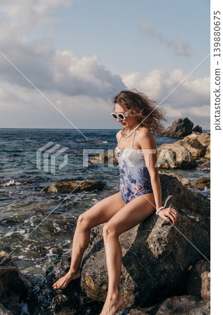 Woman sea rock sitting on a coastal rock wearing a floral swimsuit and sunglasses enjoying a breezy summer day by the ocean Woman sea rock sitting on a coastal rock wearing a floral swimsuit and sunglasses enjoying a breezy summer day by the ocean 139880675