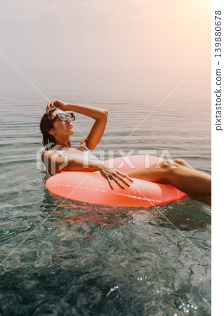 Woman floating sea young adult wearing sunglasses relaxes on a pink inflatable ring in the calm ocean water during a sunny vacation Woman floating sea young adult wearing sunglasses relaxes on a pink inflatable ring in the calm ocean water during a sunny vacation 139880678