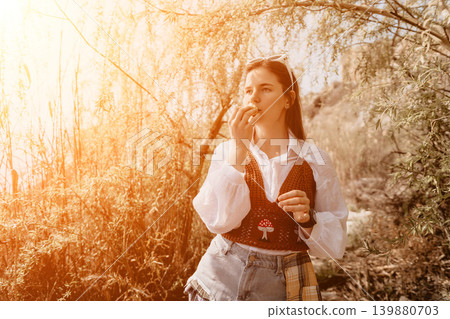 Woman eating nature. A young woman enjoys a snack while standing among autumn trees during a warm sunny excursion 139880703