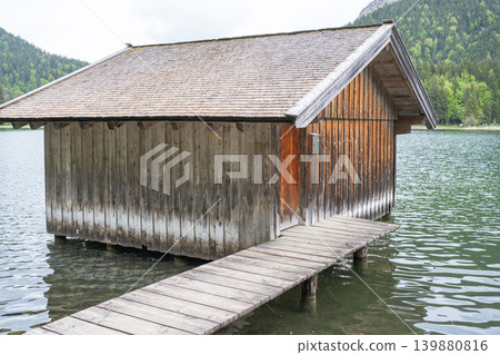 Vintage boat hut on stilts near the shore of an alpine reservoir. Concept of solitude, ecotourism, travel destinations in Europe, and serenity. Vintage boat hut on stilts near the shore of an alpine reservoir. Concept of solitude, ecotourism, travel destinations in Europe, and serenity. 139880816