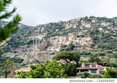 Steep, rocky mountains and tropical-style houses under a cloudy sky – on the French island of Corsica in the Mediterranean Sea. Steep, rocky mountains and tropical-style houses under a cloudy sky – on the French island of Corsica in the Mediterranean Sea. 139880999