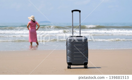 Suitcase on sandy beach in foreground while young woman in pink dress stands in calm sea water in background enjoying vacation. Holiday arrival concept Suitcase on sandy beach in foreground while young woman in pink dress stands in calm sea water in background enjoying vacation. Holiday arrival concept 139881513