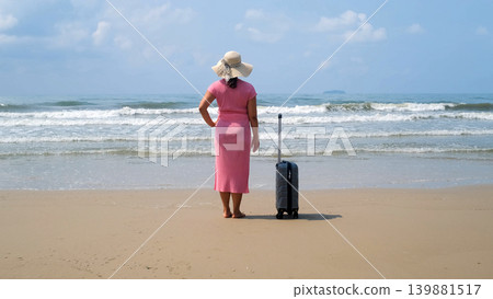 Rear view of young woman in pink dress and straw hat standing next to suitcase on beach gazing at the sea enjoying her vacation. Relaxation concept Rear view of young woman in pink dress and straw hat standing next to suitcase on beach gazing at the sea enjoying her vacation. Relaxation concept 139881517