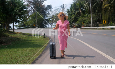 Happy young woman in straw hat walking along a road pulling a suitcase with tropical green vegetation in background on vacation. Travel arrival concept Happy young woman in straw hat walking along a road pulling a suitcase with tropical green vegetation in background on vacation. Travel arrival concept 139881521