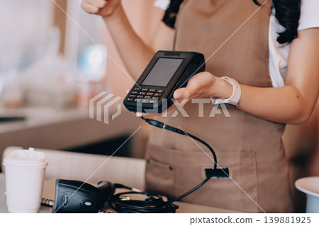 Friendly Asian female barista receives a credit card from a customer for payment. Both women are smiling in a warmly lit coffee shop with a rustic interior. Cashless payments. Small family business Friendly Asian female barista receives a credit card from a customer for payment. Both women are smiling in a warmly lit coffee shop with a rustic interior. Cashless payments. Small family business 139881925