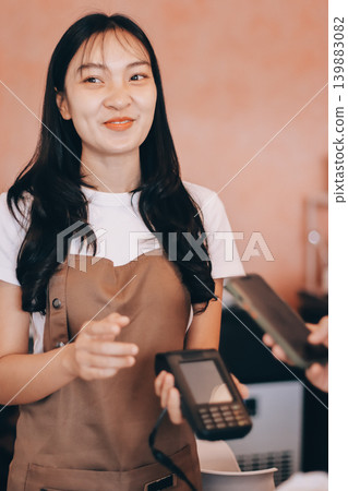Asian senior female barista adult receiving mobile payment from customer using smartphone at cafe demonstrating modern cashless transaction in cozy local family coffee shop 139883082