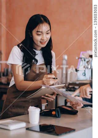 Friendly Asian female barista receives a credit card from a customer for payment. Both women are smiling in a warmly lit coffee shop with a rustic interior. Cashless payments. Small family business Friendly Asian female barista receives a credit card from a customer for payment. Both women are smiling in a warmly lit coffee shop with a rustic interior. Cashless payments. Small family business 139883083
