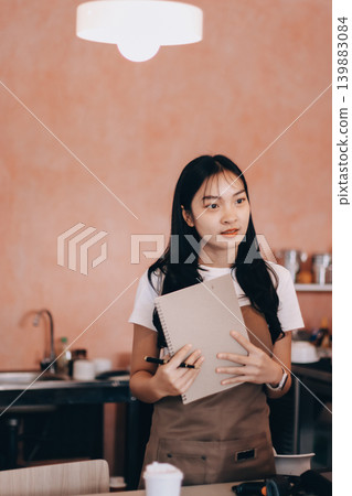 Friendly Asian female barista receives a credit card from a customer for payment. Both women are smiling in a warmly lit coffee shop with a rustic interior. Cashless payments. Small family business Friendly Asian female barista receives a credit card from a customer for payment. Both women are smiling in a warmly lit coffee shop with a rustic interior. Cashless payments. Small family business 139883084