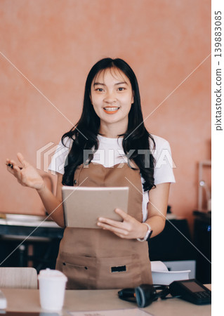 Friendly Asian female barista receives a credit card from a customer for payment. Both women are smiling in a warmly lit coffee shop with a rustic interior. Cashless payments. Small family business Friendly Asian female barista receives a credit card from a customer for payment. Both women are smiling in a warmly lit coffee shop with a rustic interior. Cashless payments. Small family business 139883085