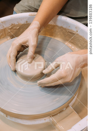 Creative sculpting. Artisan hobby. Unrecognizable female hands specialist working with raw clay pot using wheel in professional craft workspace. 139883186