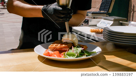 Pouring hollandaise sauce. Waffle serving. Unrecognizable professional chef man hands preparing egg benedict green salad salmon on plate in restaurant. Pouring hollandaise sauce. Waffle serving. Unrecognizable professional chef man hands preparing egg benedict green salad salmon on plate in restaurant. 139883197