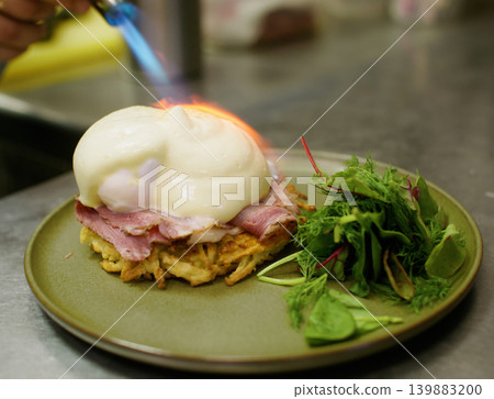 Preparing egg benedict. Healthy meal. Professional chef man hand using culinary torch burning salad ham fresh asparagus dish on plate in restaurant. Preparing egg benedict. Healthy meal. Professional chef man hand using culinary torch burning salad ham fresh asparagus dish on plate in restaurant. 139883200