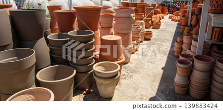 At a garden center, various terracotta flower pots and clay planters are stacked in neat rows under sunlight. Shoppers walk around, looking at the options for their gardens. At a garden center, various terracotta flower pots and clay planters are stacked in neat rows under sunlight. Shoppers walk around, looking at the options for their gardens. 139883424