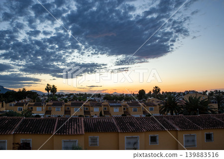 The sun sets over a Mediterranean town filled with terracotta rooftops. The sky is dramatic with clouds. This moment captures the end of a day in a lively location. 139883516