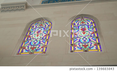 Two arched stained glass windows display colorful floral patterns. They are set in a white stone wall inside a mosque. The daylight shines through the glass. 139883843
