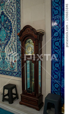 A grandfather clock made of wood is placed against a wall with blue and white floral tiles. People walk around in a mosque. Sunlight enters through windows during daytime. 139883846