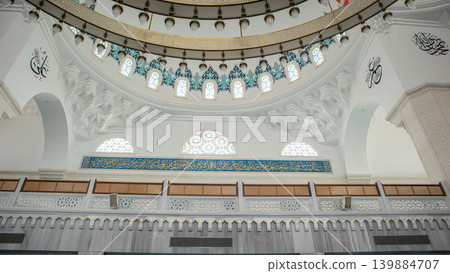 The upper balcony of a mosque features wooden details and arched designs. Golden Arabic script is displayed on a long banner. Sunlight enters from large windows above. The upper balcony of a mosque features wooden details and arched designs. Golden Arabic script is displayed on a long banner. Sunlight enters from large windows above. 139884707