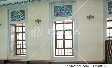 A series of arched windows can be seen in a modern mosque. The upper part shows blue floral mosaics while classic metal lattice work frames the windows. Light enters through the designs. A series of arched windows can be seen in a modern mosque. The upper part shows blue floral mosaics while classic metal lattice work frames the windows. Light enters through the designs. 139884889