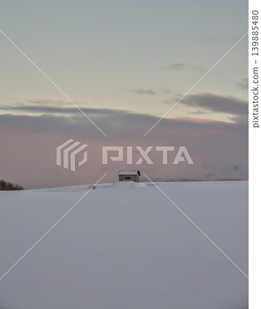 A small cabin stands alone in a vast snowfield against a pastel pink sunset sky. A small cabin stands alone in a vast snowfield against a pastel pink sunset sky. 139885480