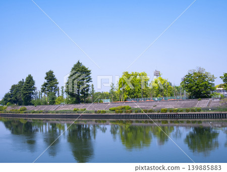 藍天下,一排排綠葉蔥蘢的樹木倒映在河面上,景色宜人。 藍天下,一排排綠葉蔥蘢的樹木倒映在河面上,景色宜人。 139885883
