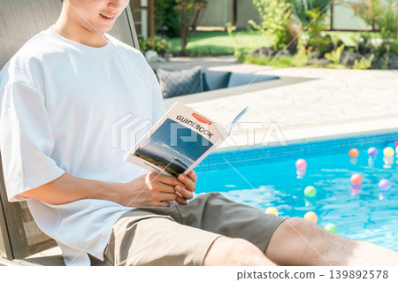 一位年輕的亞洲男子坐在泳池邊的躺椅上閱讀旅遊指南 139892578