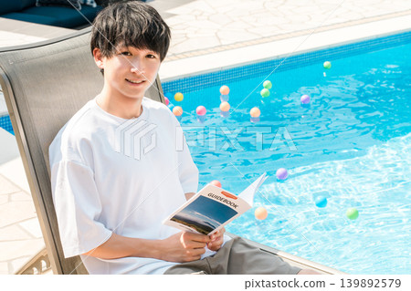 一位年輕的亞洲男子坐在泳池邊的躺椅上閱讀旅遊指南 139892579