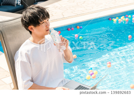 年輕的亞洲男子坐在泳池邊的躺椅上補充水分 139892683