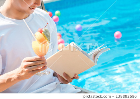 年輕的亞洲男子坐在泳池邊的躺椅上喝熱帶果汁並閱讀 139892686
