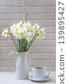 Bouquet of white daffodil flowers in white jug and white cup of tea or coffee on a whtie table against brick wall 139895427