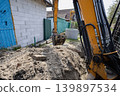 A digger is lifting dirt at a construction site next to a building. Workers are preparing the area for future work. The sun is shining overhead 139897534