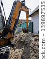 A digger works in a backyard, creating a trench for construction. Soft earth is piled high, and nearby structures are visible under a clear sky 139897535
