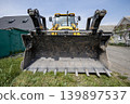 A large yellow loader is parked on a construction site with grass and buildings visible under a bright blue sky during daytime 139897537