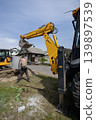 A worker is using a backhoe to dig near a building. Another machine is parked nearby. The sun is shining and the scene shows construction activity 139897539