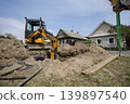 A machine is digging a deep hole in the ground next to two houses. A worker is nearby, taking part in the construction work during daytime 139897540