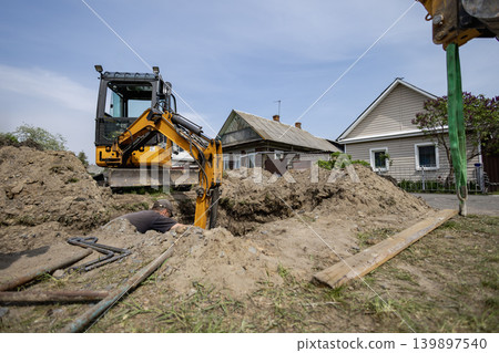 A machine is digging a deep hole in the ground next to two houses. A worker is nearby, taking part in the construction work during daytime A machine is digging a deep hole in the ground next to two houses. A worker is nearby, taking part in the construction work during daytime 139897540