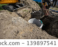 A construction crew is digging a hole to install a sewer system. A machine is lifting a concrete pipe at a site. The sun is shining in the afternoon 139897541