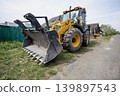 A large construction machine sits on the side of a dirt road close to houses. The machine has a bucket in front and yellow paint on its body. loader 139897543