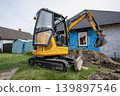A small excavator is digging in front of a house. The background shows a blue tarp on the wall and a small shed nearby. Grass surrounds the area 139897546