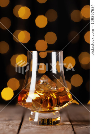Whiskey glass with amber liquid and ice rocks on an old wooden table, featuring dark background with festive golden bokeh lights, depicting luxurious celebration or premium bar setting Whiskey glass with amber liquid and ice rocks on an old wooden table, featuring dark background with festive golden bokeh lights, depicting luxurious celebration or premium bar setting 139897684